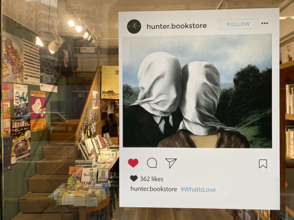 A large poster featuring two people with white cloth over their heads stands in the foreground of a bookstore.