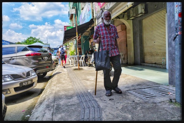 An old man walks down a sunny street with a series of shops on his left