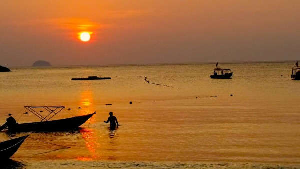 An orange sun hangs low in the air over the sea. In the foreground, a man wades through water towards a small boat